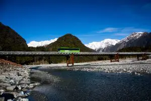 An image of a Kiwi bus crosses a scenic bridge spanning a wide river, with urban skyline and lush greenery visible in the background.