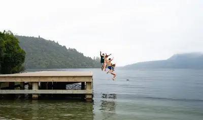 A lively group of friends joyfully jumping off a wooden jetty into a sparkling lake under a sunny sky, enjoying summer adventure.