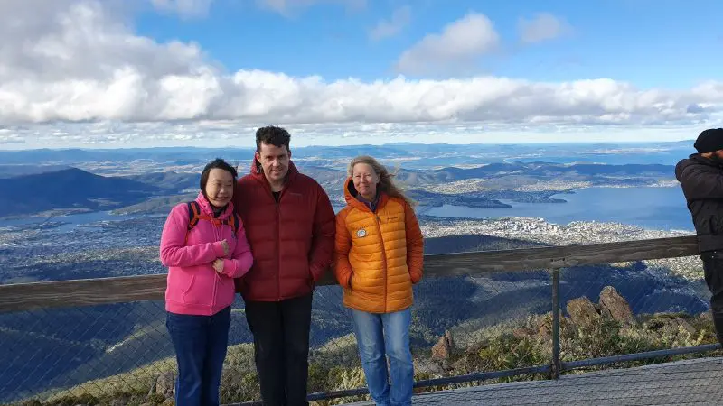 Three travellers in vibrant jackets smile on a scenic lookout deck at Gordon Dam during a Lake Pedder Wilderness Small Group Day Tour.