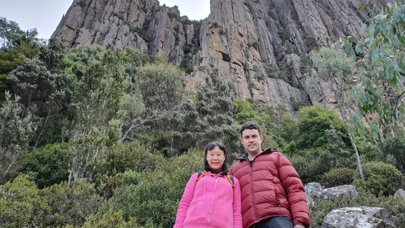 Two hikers smile at the camera on a Mount Wellington Morning Walking Tour, framed by scenic rocky cliffs and lush green trees.