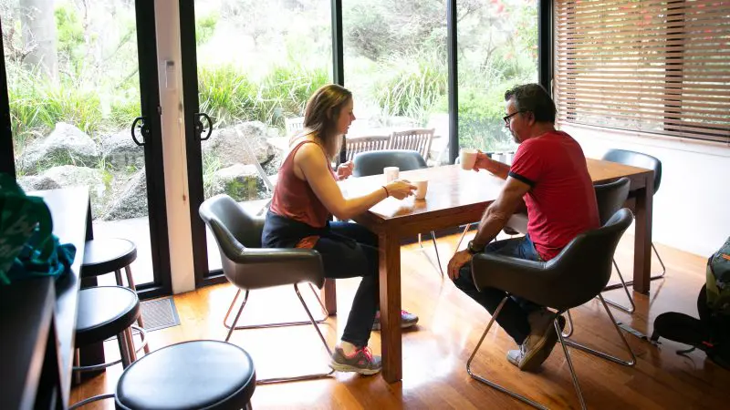 Two travellers sipping coffee by a lush garden view, discussing their Great Ocean Road and Beyond 4 Day 3 Night itinerary in detail.