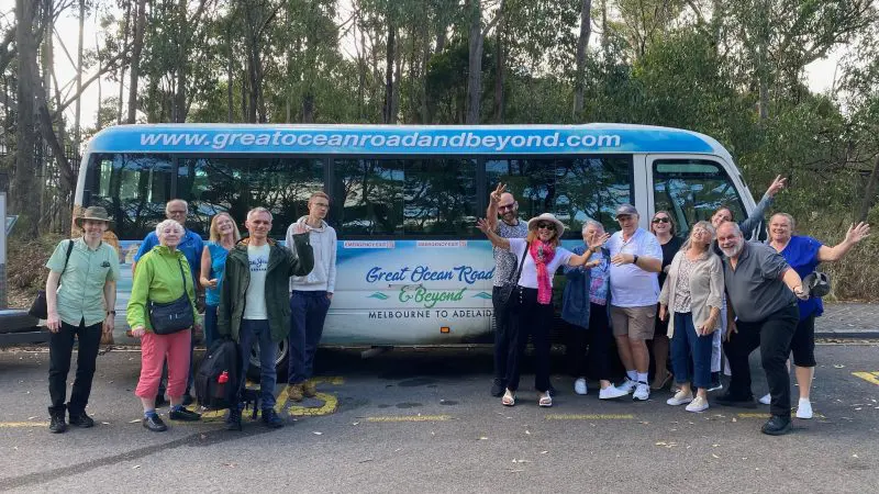 Smiling group poses by a vibrant blue Great Ocean Road And Beyond 4 Day 3 Night Tour coach, ready for an unforgettable adventure.