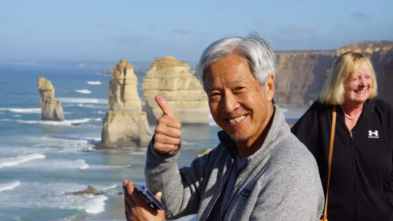Happy man giving a thumbs up in front of iconic Great Ocean Road rock formations on 4-day, 3-night guided tour adventure.