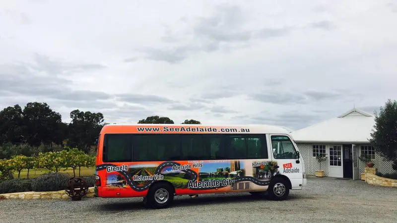 Adelaide wine tour coach parked on gravel beside lush vineyard and white winery, under dramatic clouds, perfect for wine tasting tours.