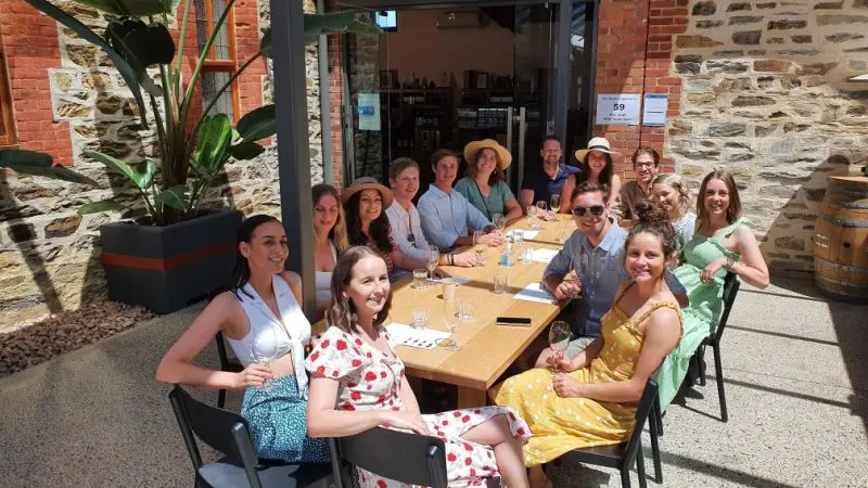 Happy group on a summer wine tasting coast tour, smiling at a sunny café table, enjoying premium wines and stunning seaside views.