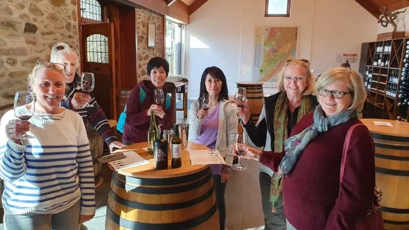 Six women at a 1 Day Wine Tasting event share smiles and clink glasses around a barrel table in a vibrant winery tasting room.