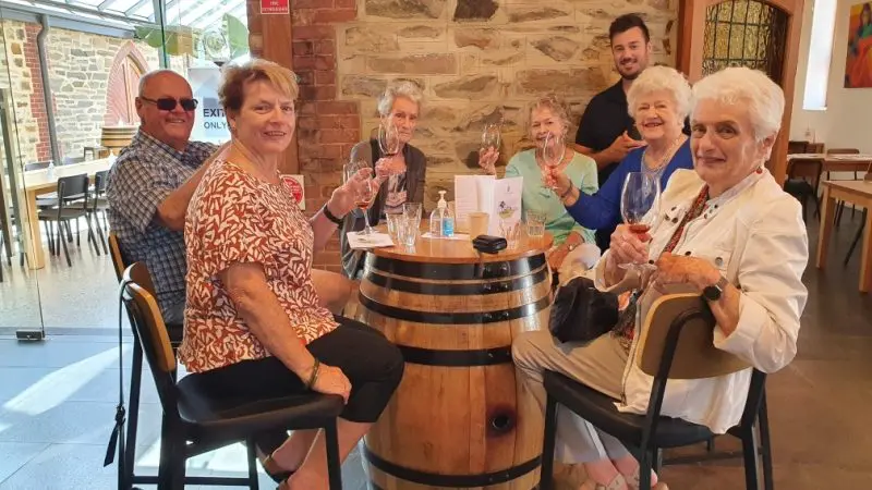 Group of six pensioners and one young man smile, toasting with wine glasses around a barrel table during an indoor Coast Afternoon Tour experience.