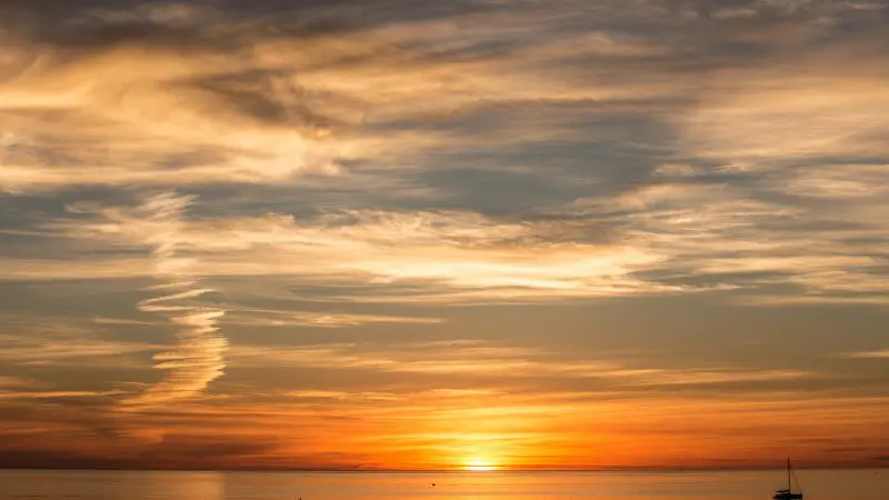Stunning orange and yellow sunset over tranquil sea, sailboat gliding by—ideal backdrop for luxury coastal wine tasting tour.