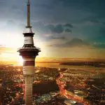 Sky Tower rising above Auckland City at sunset, vibrant lights illuminating streets and waterfront, stunning panoramic cityscape view.