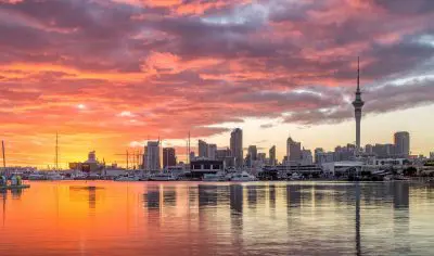 Breathtaking Auckland City skyline at sunset with vibrant colours, boats in the harbour, and city lights reflecting on shimmering water.