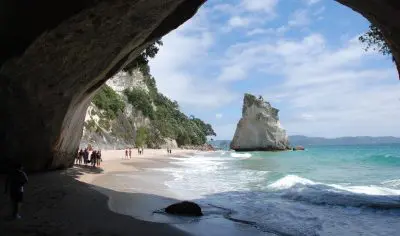 Breathtaking Coromandel coastal scene: golden sandy beach and turquoise sea framed by a rocky cave, people exploring the shore.