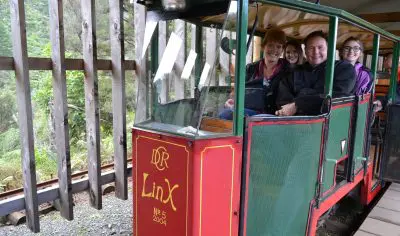 Four happy passengers enjoy a private LinX red train carriage tour at a picturesque outdoor railway station, surrounded by scenic views.