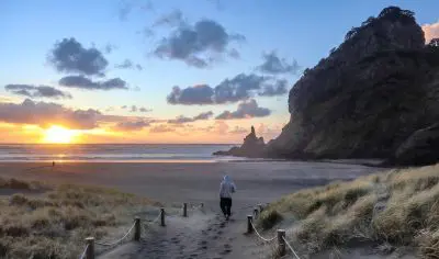 A person in a hoodie strolls along a sandy West Coast path towards the beach at sunset, showcasing stunning coastal scenery.