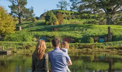 A family enjoys the scenic Hobbiton Movie Set with iconic hillside hobbit houses by a tranquil pond under a clear blue sky in New Zealand.