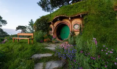 Enchanting round hobbit door nestled in a lush grassy hillside at Hobbiton Film Set, adorned with vibrant flowers, bench, and winding path.
