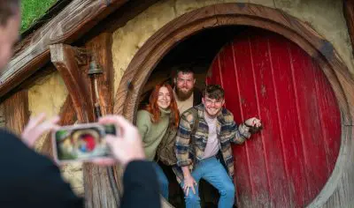 Three happy visitors pose by a red Hobbit door at the Hobbiton Movie Set as their photo is captured on a smartphone for memories.