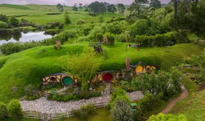 Experience the iconic Hobbiton Film Set with charming hillside hobbit homes, vibrant greenery, and a serene pond beneath a pink sky.