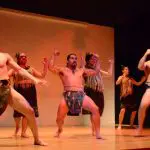 Traditional Māori performers in Auckland present an authentic haka dance in vibrant attire on a wooden stage during a Māori Experience.