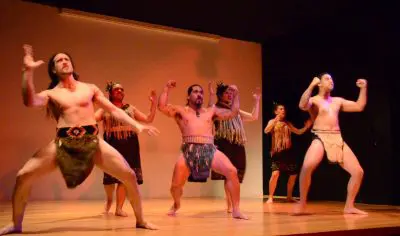 Traditional Māori performers in Auckland present an authentic haka dance in vibrant attire on a wooden stage during a Māori Experience.