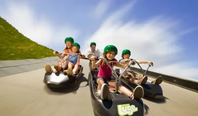 Five kids in helmets race luges downhill in Rotorua on a sunny day, laughing and enjoying thrilling outdoor adventures together.