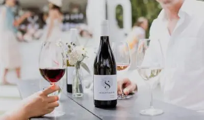 Premium red wine bottle and glasses on an outdoor table in a scenic Wine Village, with two people and a floral vase centrepiece.
