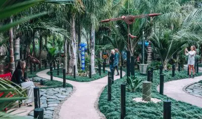 Visitors on a private tour capture photos amid lush gardens filled with vibrant palm trees, artistic sculptures, and winding stone paths.