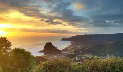 Stunning golden sunset at Piha Beach highlighting dramatic rugged coastline, iconic Lion Rock, sandy shore, and lush green hills.