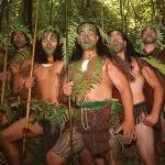 Six Māori warriors with intricate tattoos and spears stand in a lush forest, set to welcome guests to an immersive Māori Dinner Show.