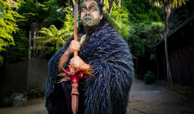 Individual dressed in authentic Māori clothing holding a carved wooden staff at a Māori cultural show, surrounded by vibrant greenery.