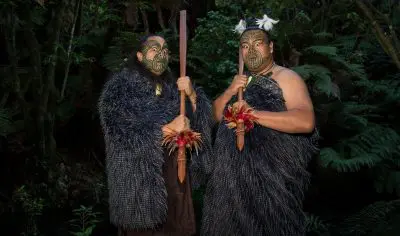 Experience an exclusive private tour with two individuals in authentic Māori dress and facial moko holding carved staffs in a lush forest.