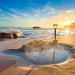A spade in wet sand by a water-filled hole catches sunrise on Coromandel Beach as ocean waves crash, capturing New Zealand’s beauty.