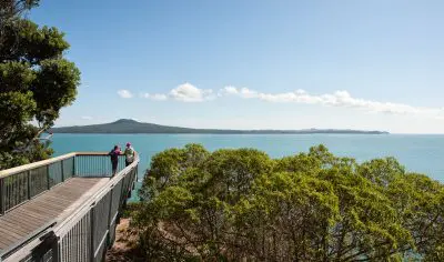 Visitors enjoy panoramic views from a wooden walkway overlooking Auckland city skyline and scenic islands beneath a clear blue sky.