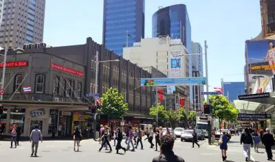 Pedestrians cross a bustling Auckland city street amid modern buildings and retail shops under clear blue skies and brilliant sunshine.