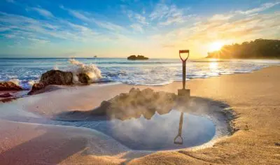 A metal spade in golden sand beside a steaming geothermal pool at Hot Water Beach, New Zealand, with waves and rocky shoreline behind.
