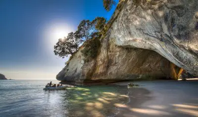 A small boat approaches a sunlit cave at Cathedral Cove, gliding through turquoise waters beside dramatic Coromandel Peninsula cliffs.