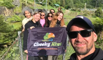 Group of happy travellers on a bridge holding a Cheeky Kiwi Tours flag, set for a Coromandel adventure, blue sky in the background.