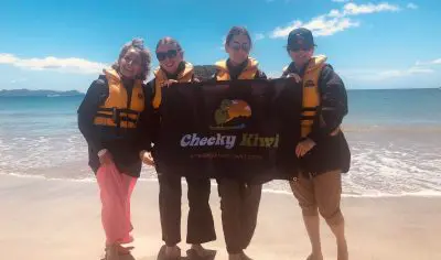 Four smiling people in life jackets stand on a sunny Cathedral Cove beach, holding a Cheeky Kiwi Travel banner for an adventure tour.