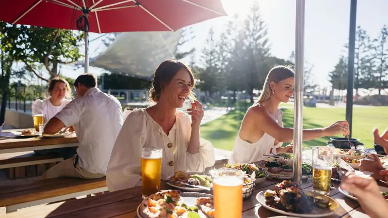 Group of women laughing and sharing gourmet food and drinks outdoors at a lively Margaret River Wildlife Adventure restaurant experience.