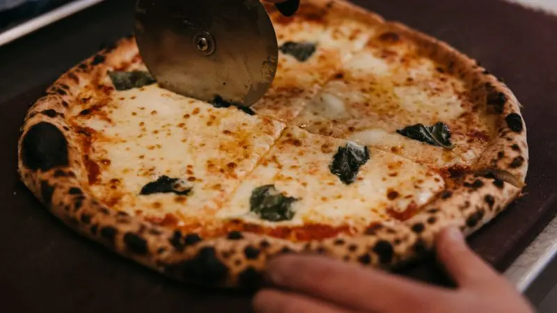 Close-up of a hand slicing a cheesy basil pizza with a cutter—ideal meal after exploring Margaret River and its famous wildlife.