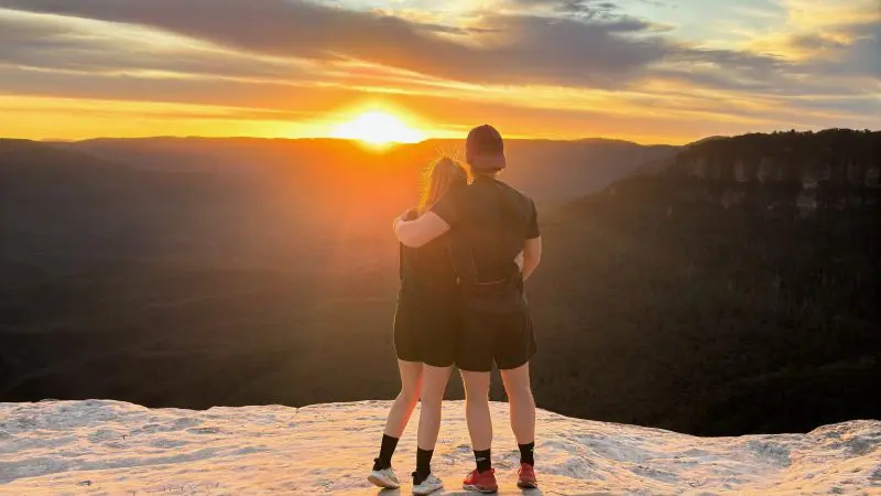 Romantic couple embraces on a rocky Blue Mountains cliff at sunset during a 1 Day Wilderness Tour, overlooking scenic valleys.