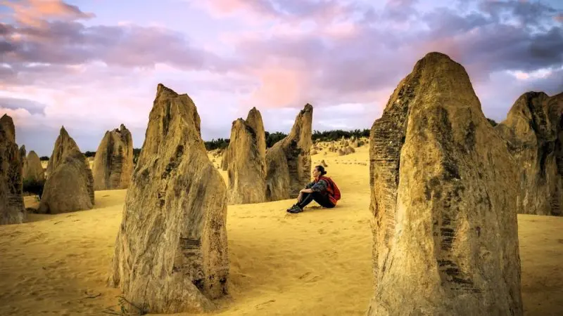 Experience the awe of the Pinnacles Desert as a traveller relaxes on golden sands beside striking limestone pillars at sunset with Autopia.