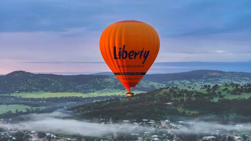 A bright orange hot air balloon labelled “Liberty” soars above Perth’s scenic Avon Valley during a gourmet balloon flight with breakfast.