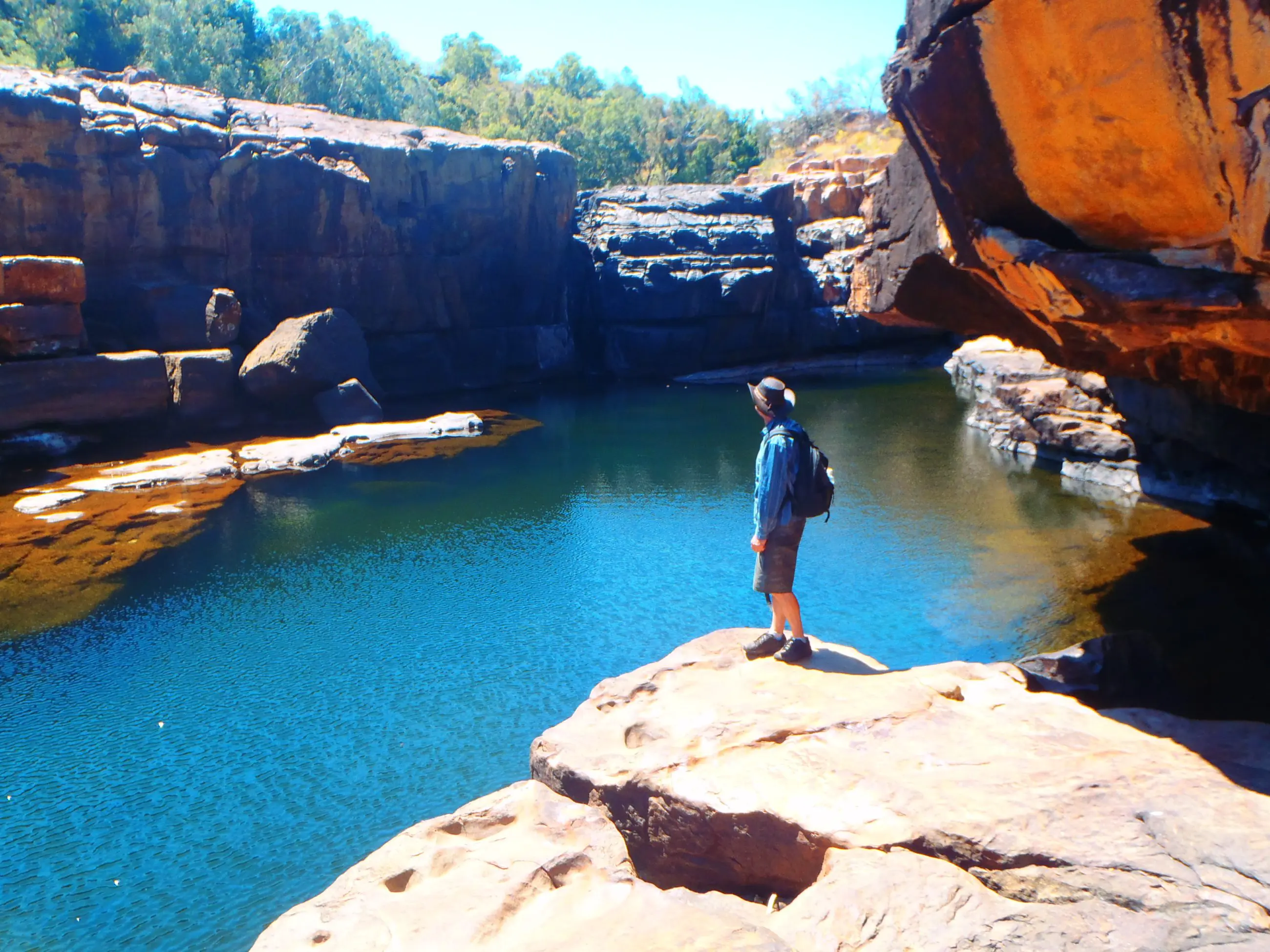 Adventurer with rucksack stands on rocky ledge during Kakadu Koolpin Tour, gazing over a pristine blue river and lush natural landscape.