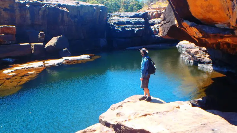 Adventurer with rucksack stands on rocky ledge during Kakadu Koolpin Tour, gazing over a pristine blue river and lush natural landscape.