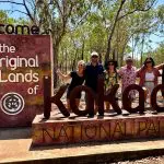 Group of six happy campers by the Welcome to Kakadu sign during a 3 Day Kakadu Adventure, June–Sept, in bright, wooded outback.