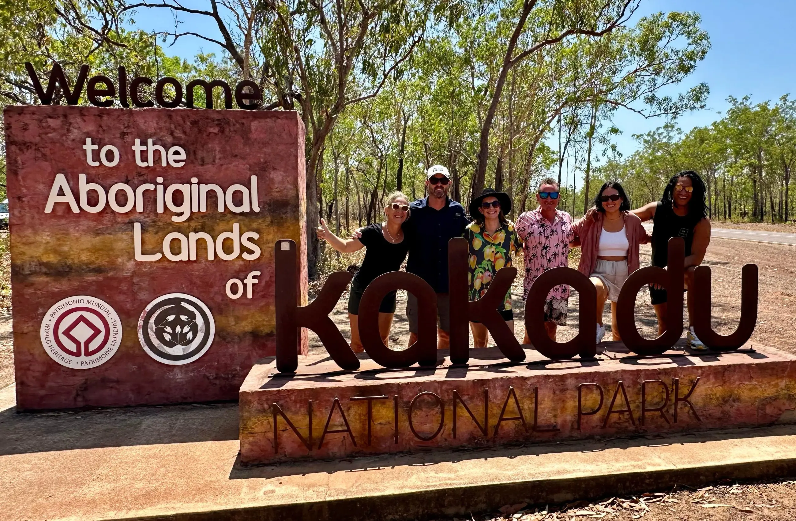 Group of six tourists smiling at the Kakadu National Park sign, eager to discover top Kakadu Tour Highlights amid lush greenery.