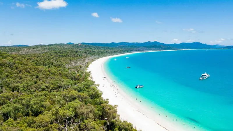 Stunning aerial view of Whitehaven Beach with crystal-clear turquoise waters, verdant forest, boats anchored near shore, and sunny skies.