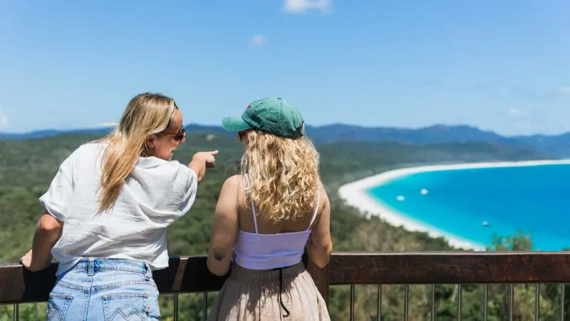 Two women admire the panoramic view at Whitehaven Beach, with one pointing towards crystal-clear turquoise waters under a pristine blue sky.