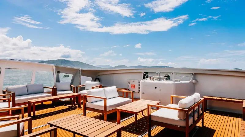 Luxurious yacht deck with plush seating, stylish wooden tables, and panoramic Whitehaven Beach views—sun-soaked relaxation all day.