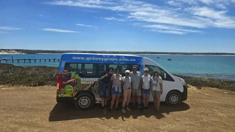 Group of six travellers smiles beside a vibrant tour van on the scenic Kangaroo Island coast during a 2-day wildlife adventure tour.